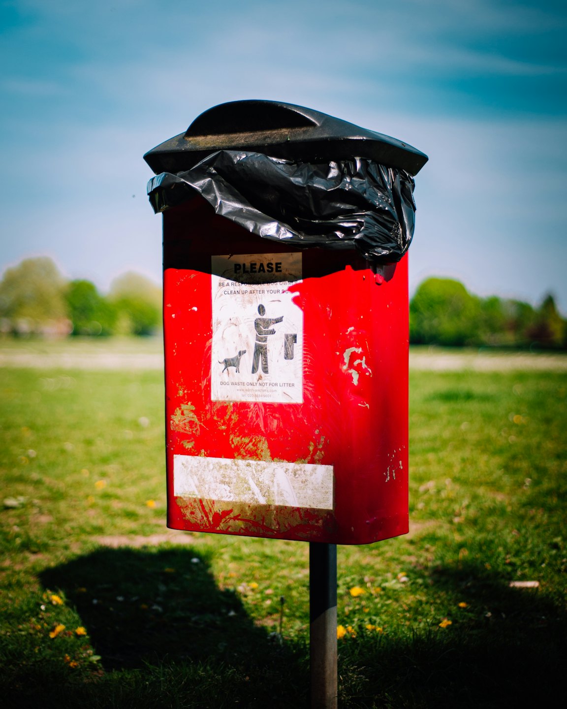A weathered red dog waste bin stands in a grassy field.